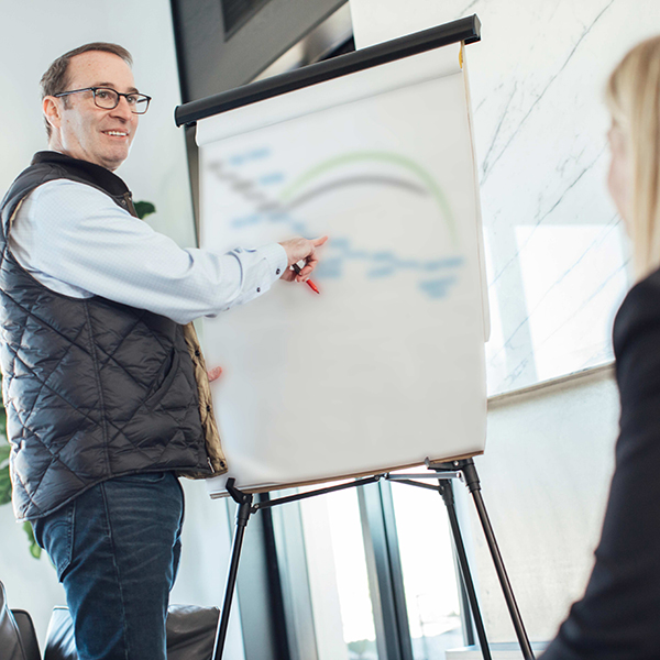 strategic sale - a woman standing at a whiteboard in an office talking to a man who is sitting in front of her